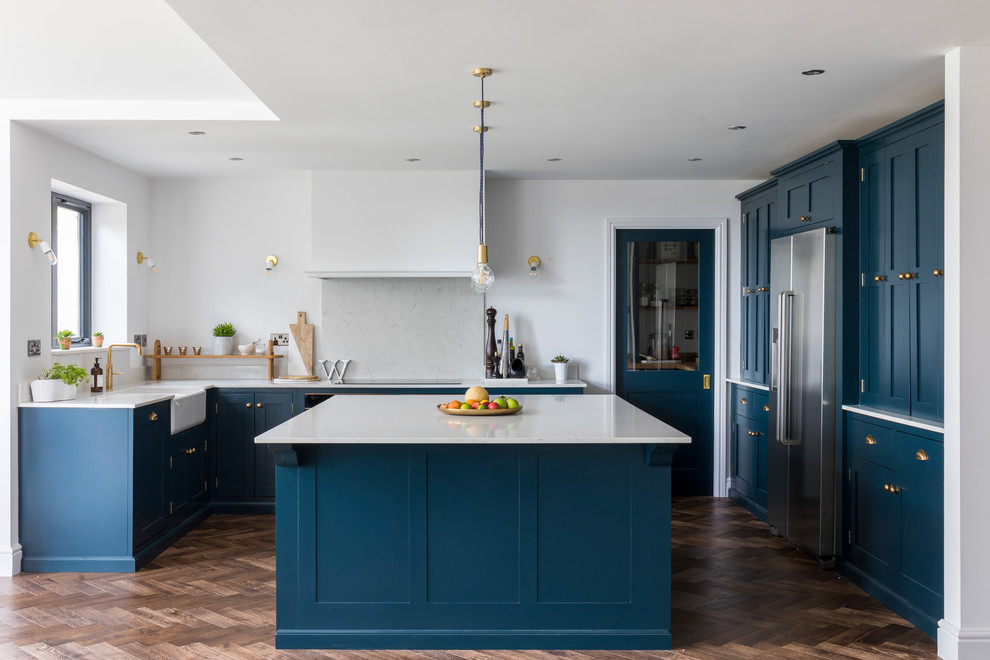 Photo of a mid-sized transitional u-shaped eat-in kitchen in London with a farmhouse sink, shaker cabinets, blue cabinets, grey splashback, stainless steel appliances, dark hardwood floors, with island, brown floor and white benchtop.