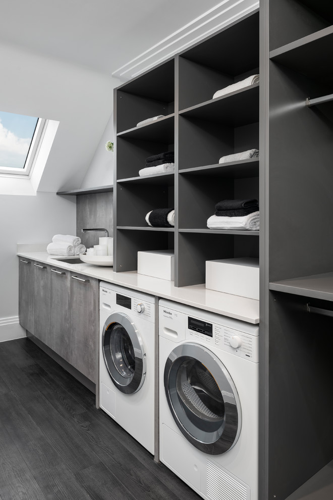 Photo of a large contemporary single-wall dedicated laundry room in Buckinghamshire with an undermount sink, open cabinets, grey cabinets, white walls, dark hardwood floors, a side-by-side washer and dryer and white benchtop.