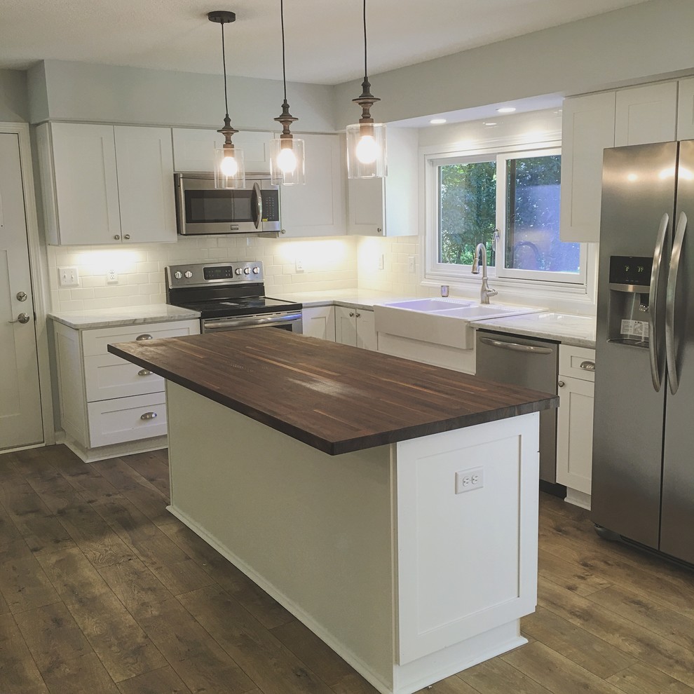 Photo of a mid-sized country l-shaped eat-in kitchen in Minneapolis with a farmhouse sink, shaker cabinets, white cabinets, wood benchtops, white splashback, ceramic splashback, stainless steel appliances, laminate floors, with island, brown floor and brown benchtop.