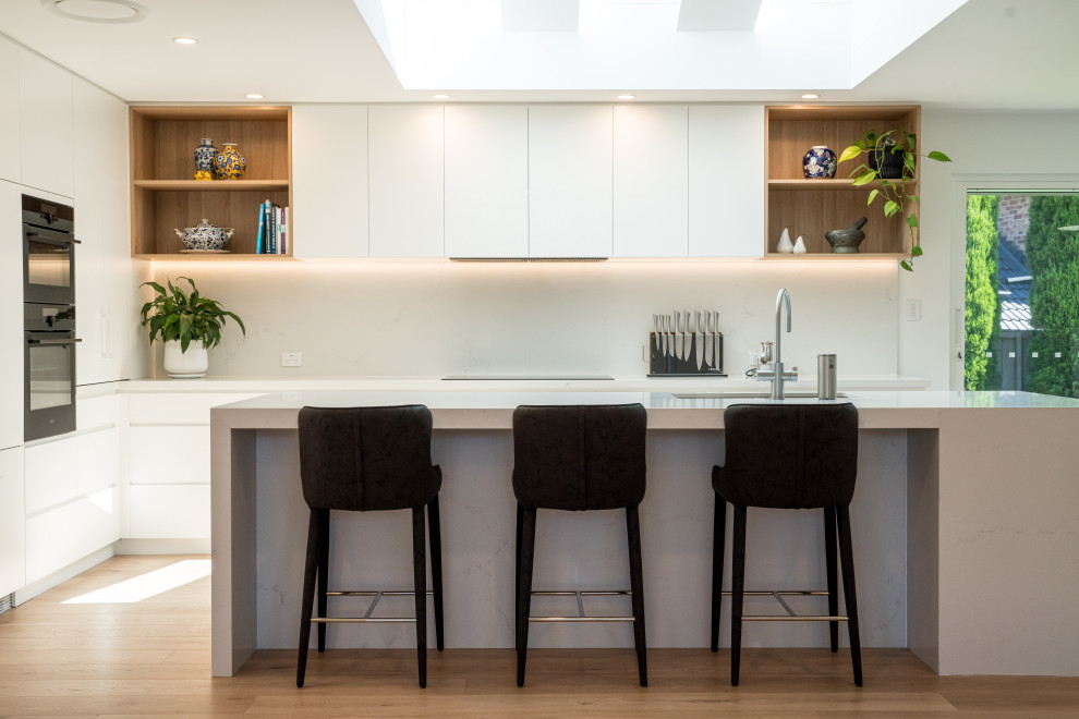 Photo of a large modern l-shaped open plan kitchen in Sydney with a double-bowl sink, flat-panel cabinets, white cabinets, quartz benchtops, white splashback, engineered quartz splashback, black appliances, laminate floors, with island, brown floor and white benchtop.