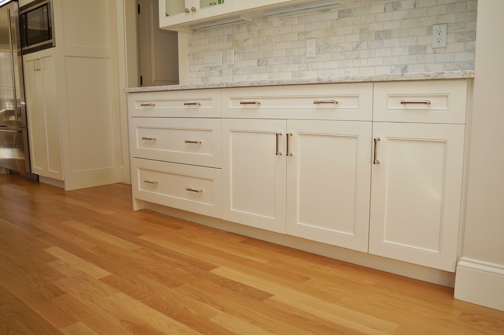 Example of a large transitional l-shaped light wood floor and brown floor kitchen pantry design in Boston with an undermount sink, recessed-panel cabinets, white cabinets, solid surface countertops, gray backsplash, stone tile backsplash, stainless steel appliances, an island and white countertops