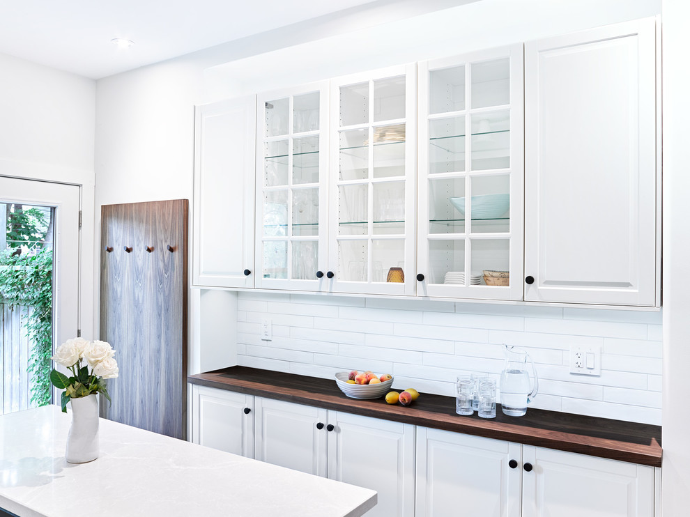 Example of a mid-sized transitional galley porcelain tile and gray floor enclosed kitchen design in Toronto with an undermount sink, recessed-panel cabinets, white cabinets, white backsplash, subway tile backsplash, stainless steel appliances, an island and wood countertops