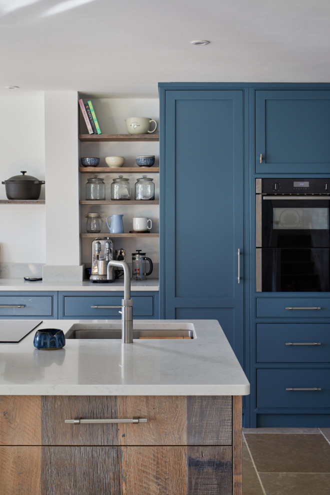 Photo of a large modern l-shaped open plan kitchen in London with a farmhouse sink, shaker cabinets, blue cabinets, quartzite benchtops, white splashback, window splashback, panelled appliances, ceramic floors, with island, brown floor and white benchtop.