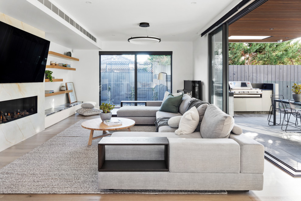 This is an example of a large modern open concept family room in Melbourne with white walls, light hardwood floors, a standard fireplace, a plaster fireplace surround, a wall-mounted tv and beige floor.