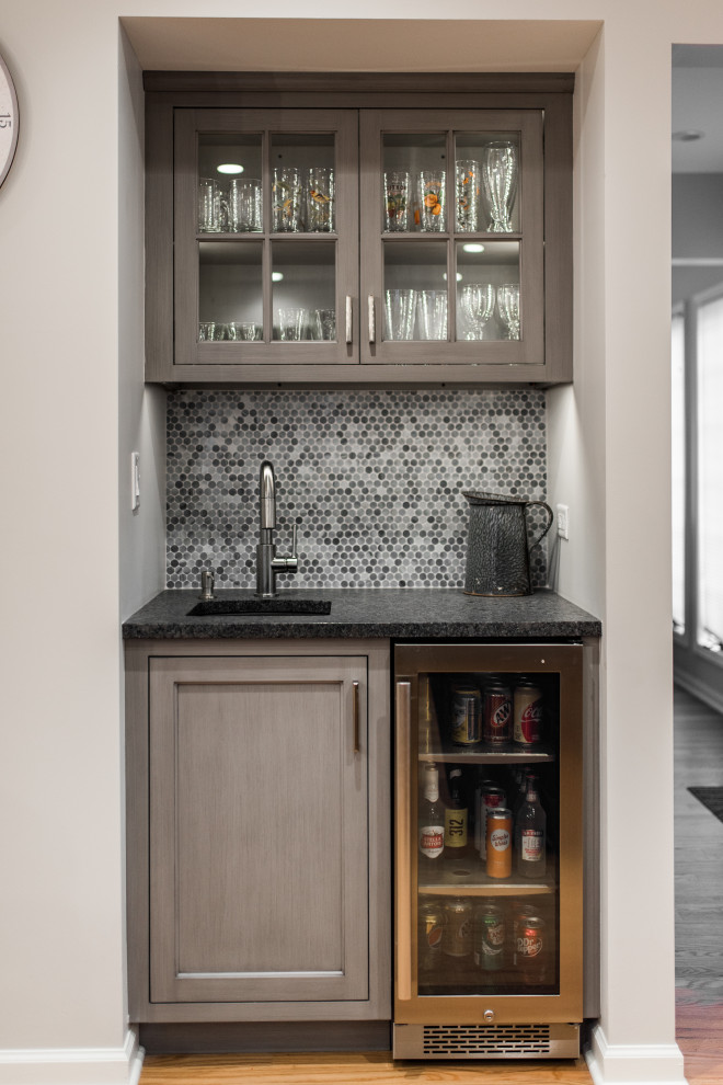 This is an example of a small transitional wet bar in Chicago with an undermount sink, glass-front cabinets, medium wood cabinets, grey splashback, brown floor and black benchtop.
