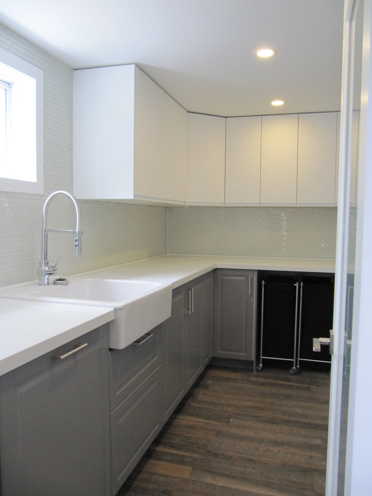 This is an example of a mid-sized modern l-shaped utility room in Toronto with a farmhouse sink, flat-panel cabinets, grey cabinets, quartzite benchtops, white walls, vinyl floors, a stacked washer and dryer, brown floor and white benchtop.
