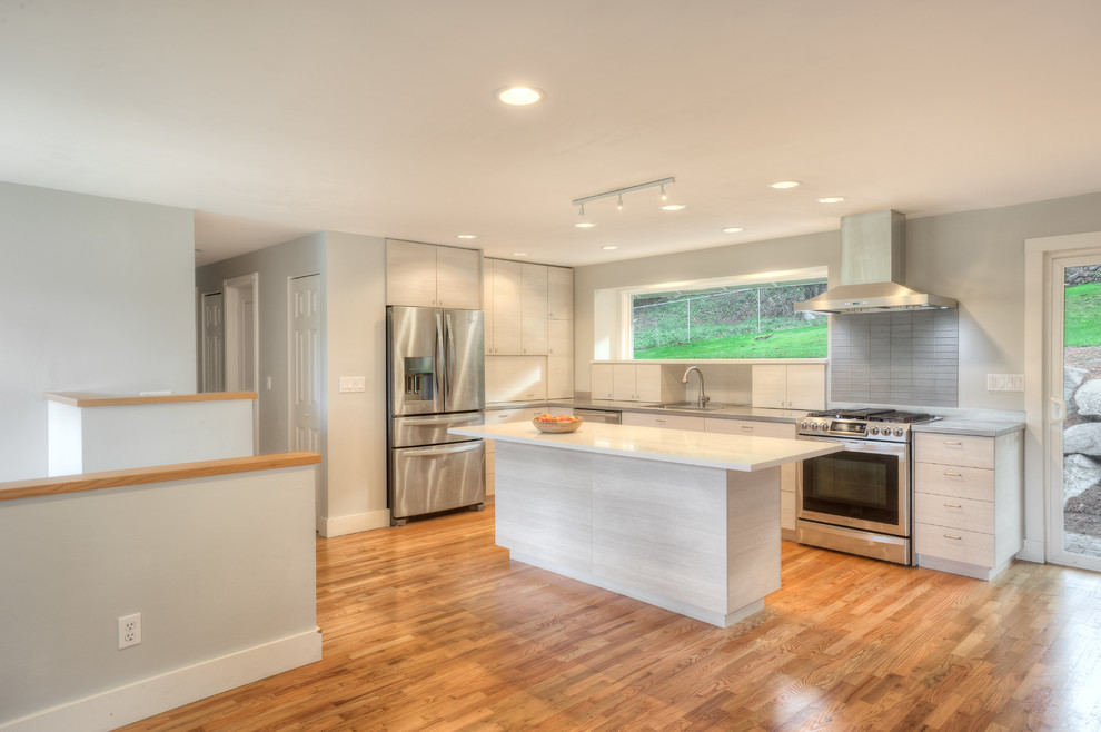 Small contemporary l-shaped open plan kitchen in Seattle with a single-bowl sink, flat-panel cabinets, stainless steel benchtops, grey splashback, ceramic splashback, stainless steel appliances, light hardwood floors, with island and white cabinets.
