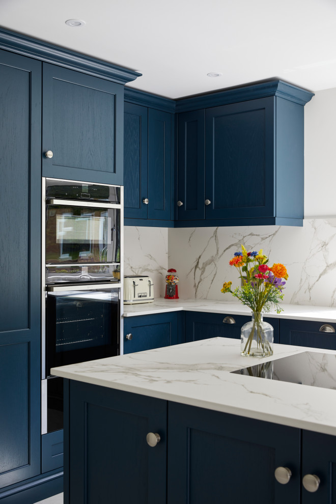 This is an example of a large contemporary l-shaped kitchen/diner in London with a belfast sink, recessed-panel cabinets, blue cabinets, engineered stone countertops, white splashback, stone slab splashback, stainless steel appliances, an island and white worktops.