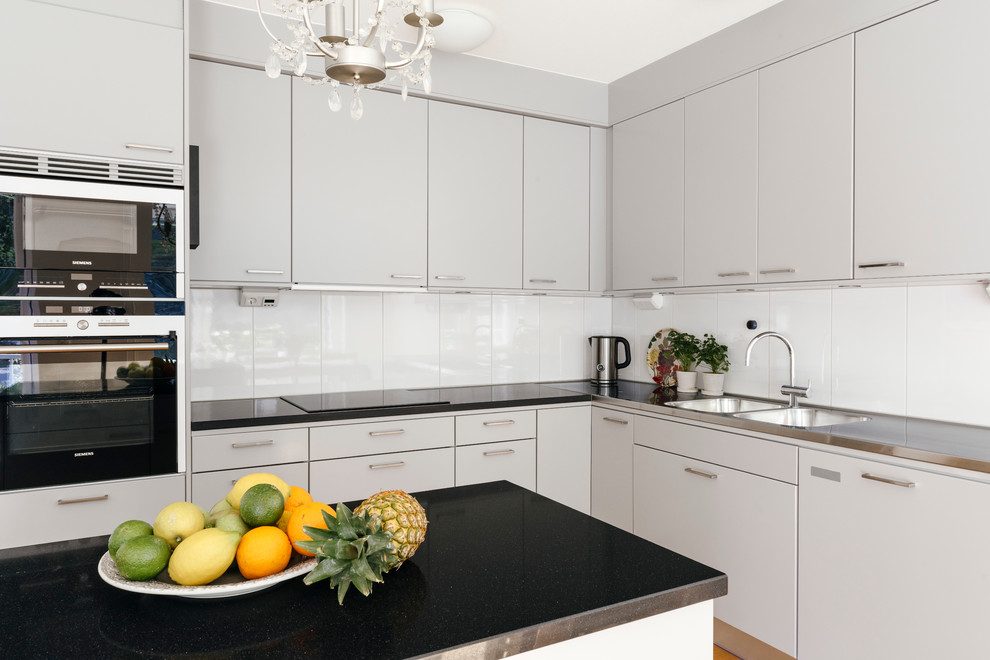 Design ideas for a mid-sized contemporary l-shaped kitchen in Stockholm with a double-bowl sink, flat-panel cabinets, white cabinets, glass tile splashback, black appliances, with island, stainless steel benchtops and white splashback.