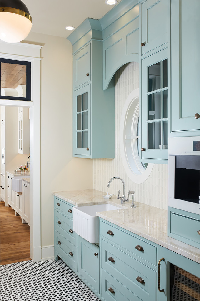 This is an example of a mid-sized beach style single-wall kitchen pantry with a farmhouse sink, glass-front cabinets, beige cabinets, marble benchtops, white splashback, window splashback, white appliances, porcelain floors, no island and multi-coloured floor.