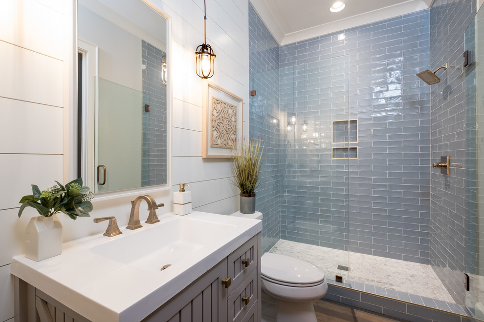 Design ideas for a mid-sized country bathroom in Charlotte with grey cabinets, a two-piece toilet, blue tile, glass tile, beige walls, medium hardwood floors, an undermount sink, brown floor, a hinged shower door, white benchtops and shaker cabinets.