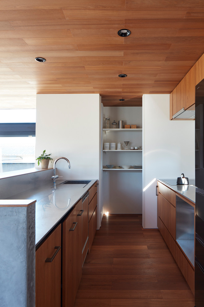 Asian galley kitchen in Other with an integrated sink, flat-panel cabinets, medium wood cabinets, stainless steel benchtops, black appliances, medium hardwood floors, a peninsula and brown floor.