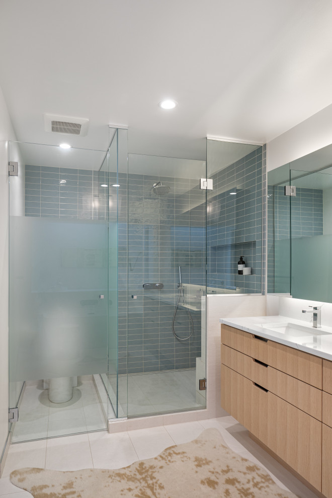Bild på ett funkis vit vitt badrum, med släta luckor, skåp i ljust trä, en hörndusch, blå kakel, vita väggar, ett undermonterad handfat, vitt golv och dusch med gångjärnsdörr