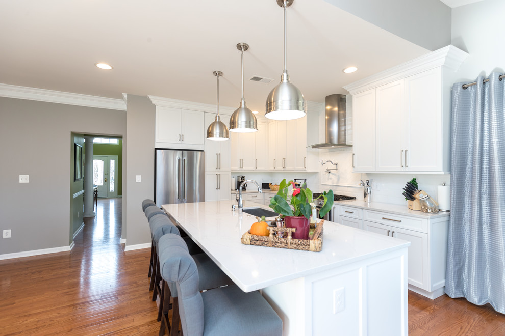 Transitional l-shaped medium tone wood floor and brown floor open concept kitchen photo in DC Metro with an undermount sink, recessed-panel cabinets, white cabinets, white backsplash, ceramic backsplash, stainless steel appliances, an island and gray countertops