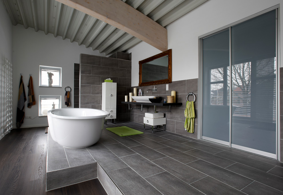 Photo of an expansive industrial master bathroom in Other with a vessel sink, a freestanding tub, flat-panel cabinets, white cabinets, gray tile, stone tile, white walls, slate floors, a curbless shower, laminate benchtops, black benchtops, a wall-mount toilet, grey floor and an open shower.