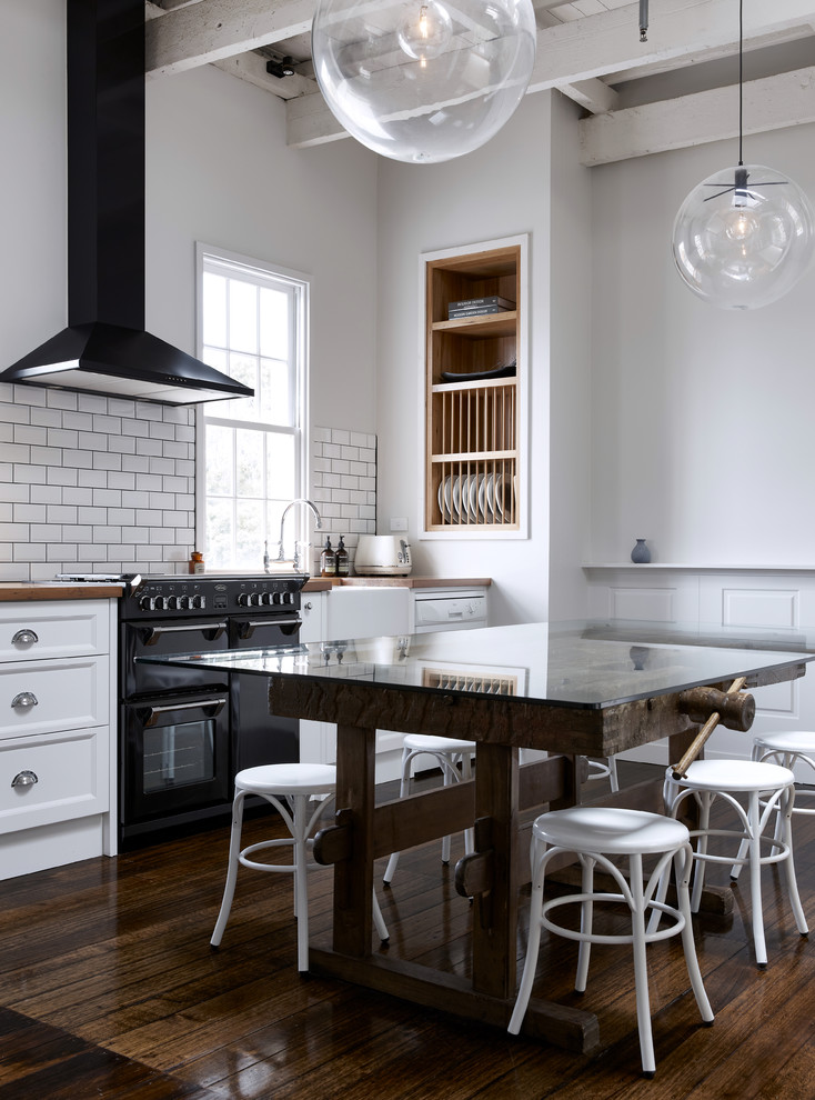 Design ideas for an industrial single-wall kitchen in Melbourne with a farmhouse sink, recessed-panel cabinets, white cabinets, white splashback, subway tile splashback, black appliances, dark hardwood floors and with island.