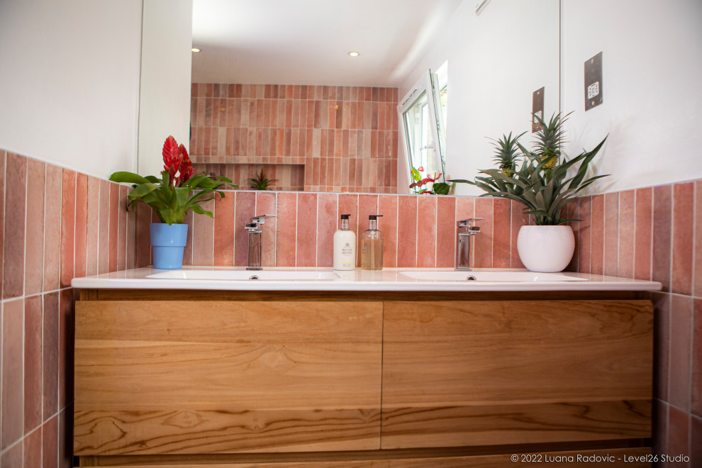 This is an example of a medium sized modern bathroom in London with flat-panel cabinets, light wood cabinets, a built-in bath, a built-in shower, a wall mounted toilet, pink tiles, porcelain tiles, white walls, ceramic flooring, a wall-mounted sink, solid surface worktops, multi-coloured floors, an open shower, a wall niche, double sinks and a freestanding vanity unit.