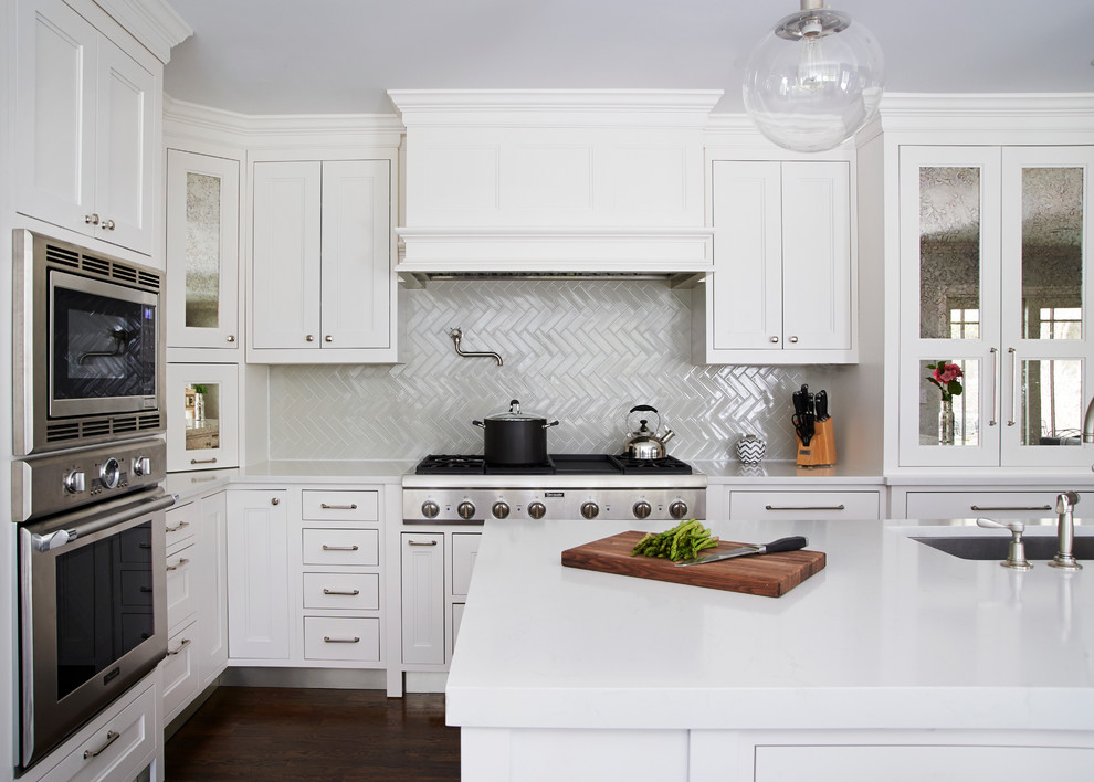 Mid-sized transitional l-shaped dark wood floor eat-in kitchen photo in New York with a farmhouse sink, shaker cabinets, white cabinets, quartz countertops, gray backsplash, ceramic backsplash, paneled appliances and an island