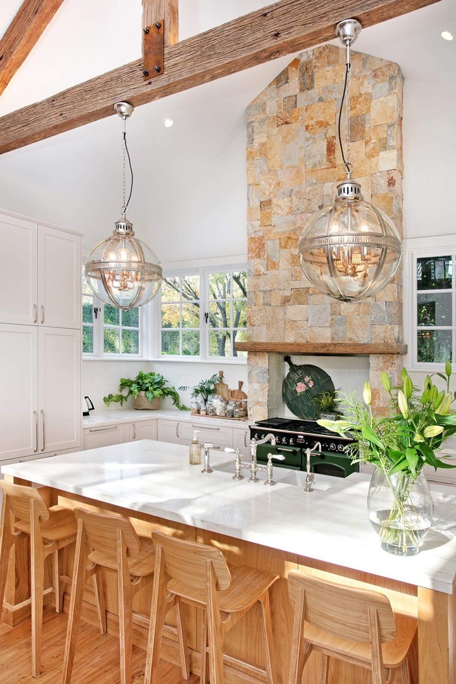 This is an example of a large country l-shaped eat-in kitchen in Sydney with a farmhouse sink, shaker cabinets, beige cabinets, solid surface benchtops, beige splashback, porcelain splashback, panelled appliances, light hardwood floors, with island and beige floor.