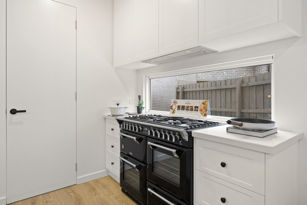 This is an example of a mid-sized transitional galley open plan kitchen in Melbourne with an undermount sink, shaker cabinets, white cabinets, quartz benchtops, window splashback, black appliances, laminate floors, with island, brown floor and white benchtop.