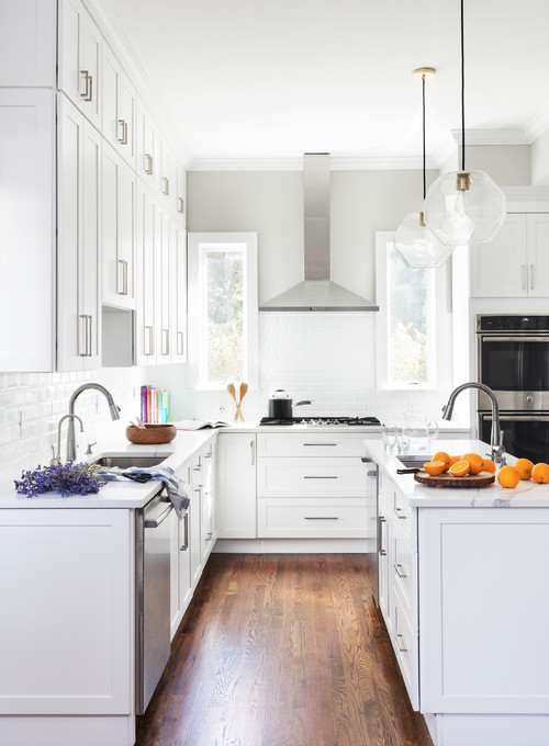Bright white U-shaped kitchen with shaker-style RTA cabinets to the ceiling, subway-tile backsplash, stainless range hood and wall oven, globe pendants, and warm wood floors.