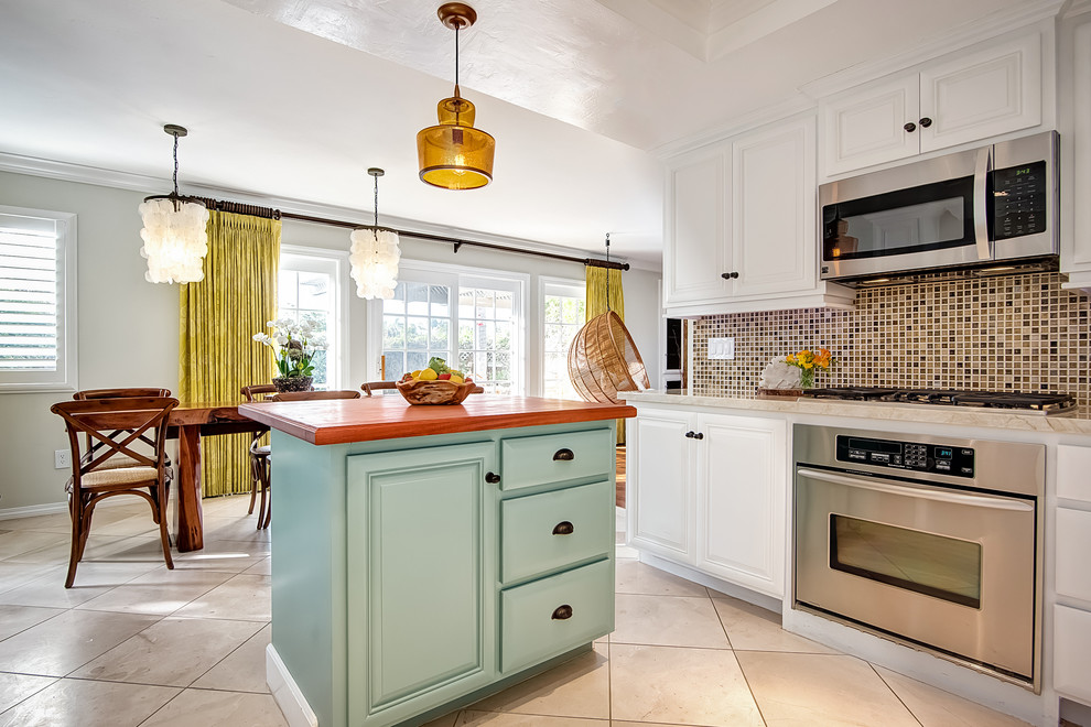 Inspiration for a mid-sized tropical u-shaped eat-in kitchen in San Diego with a farmhouse sink, beaded inset cabinets, white cabinets, onyx benchtops, multi-coloured splashback, glass tile splashback, stainless steel appliances, travertine floors and with island.