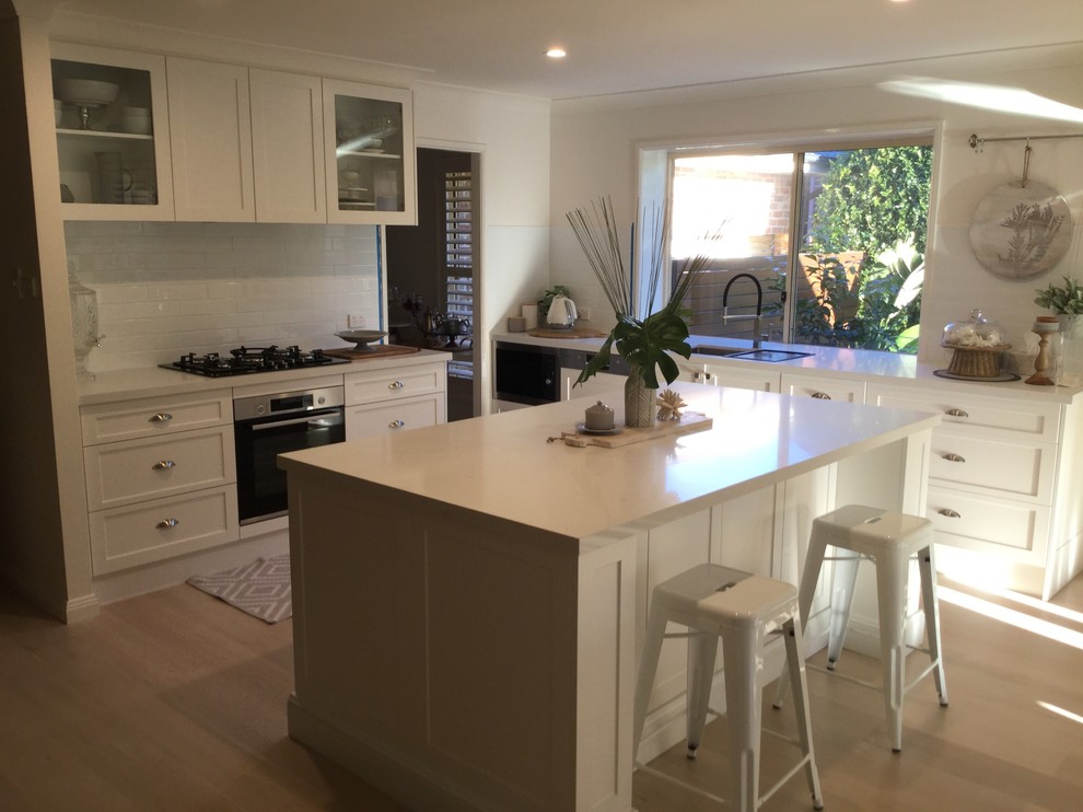 This is an example of a mid-sized transitional eat-in kitchen in Sydney with an undermount sink, shaker cabinets, white cabinets, quartz benchtops, white splashback, subway tile splashback, stainless steel appliances, light hardwood floors, with island, brown floor and white benchtop.