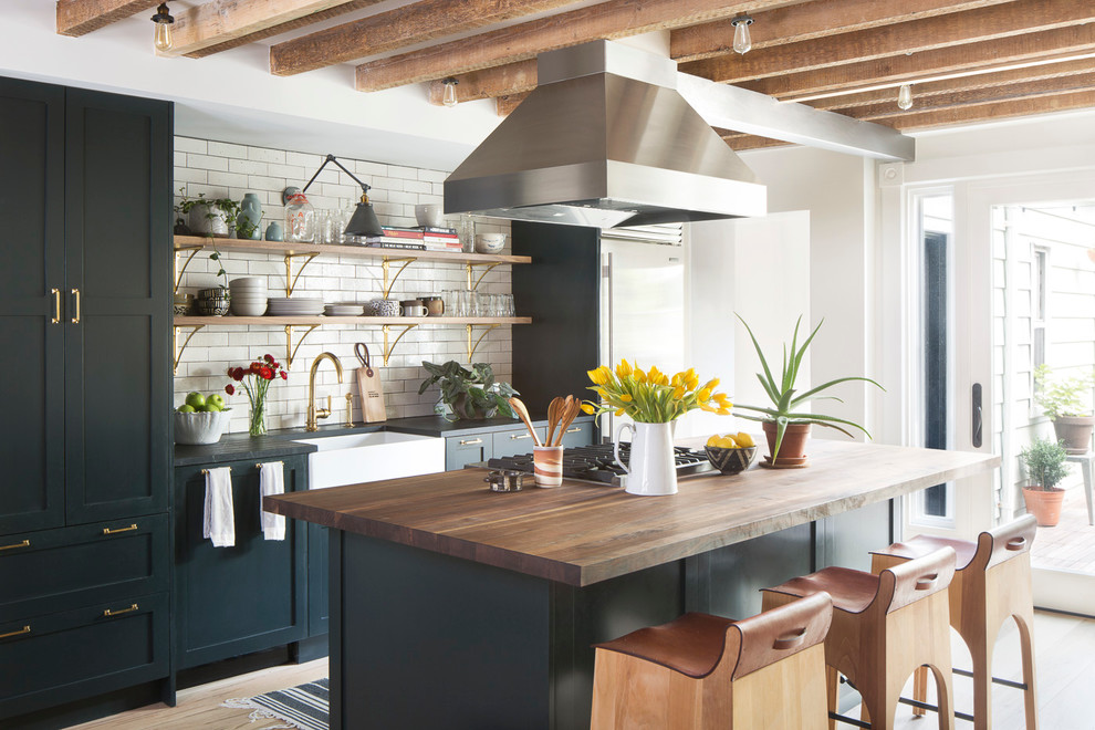Inspiration for a large transitional galley open plan kitchen in New York with a farmhouse sink, open cabinets, white splashback, subway tile splashback, stainless steel appliances, light hardwood floors, with island, black cabinets, wood benchtops and beige floor.