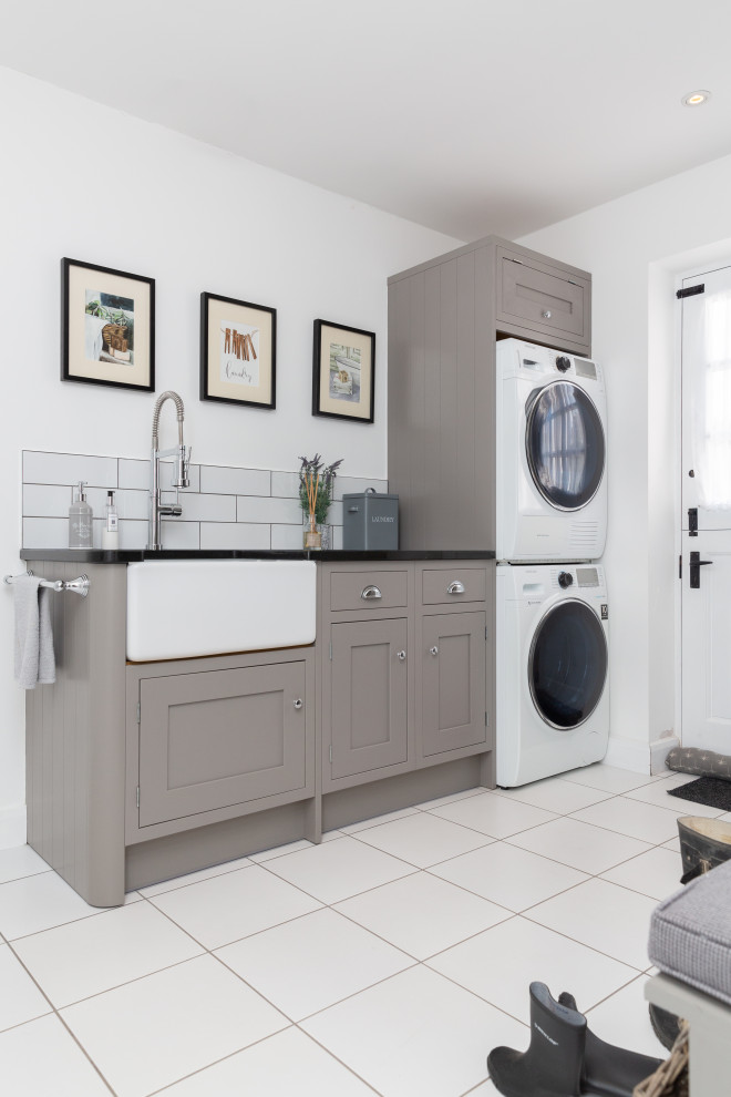 Photo of a transitional single-wall utility room in Other with a farmhouse sink, shaker cabinets, grey cabinets, subway tile splashback, white walls, a stacked washer and dryer, white floor and black benchtop.