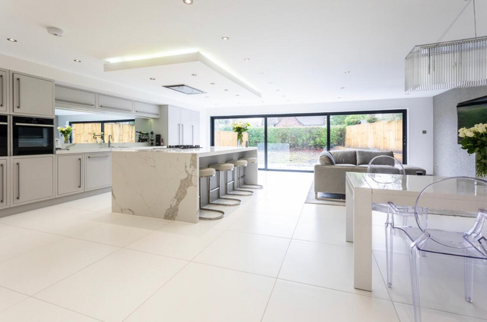 Expansive contemporary single-wall open plan kitchen in Manchester with a drop-in sink, flat-panel cabinets, grey cabinets, quartzite benchtops, metallic splashback, mirror splashback, black appliances, porcelain floors, with island, white floor and grey benchtop.