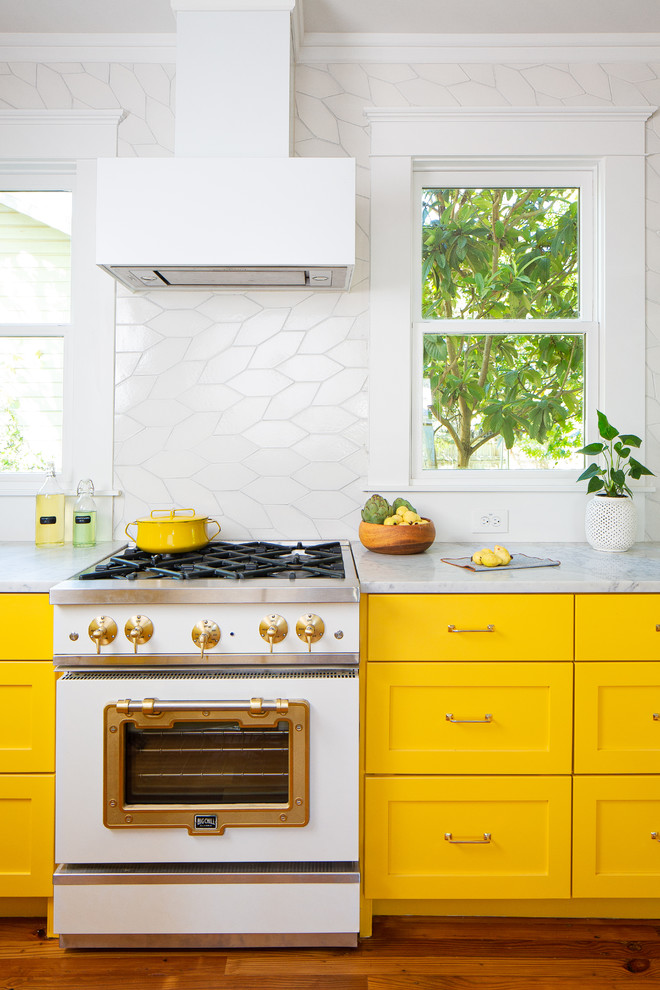Design ideas for a mid-sized eclectic l-shaped eat-in kitchen in Austin with an undermount sink, recessed-panel cabinets, yellow cabinets, marble benchtops, white splashback, ceramic splashback, white appliances, medium hardwood floors, with island, brown floor and white benchtop.