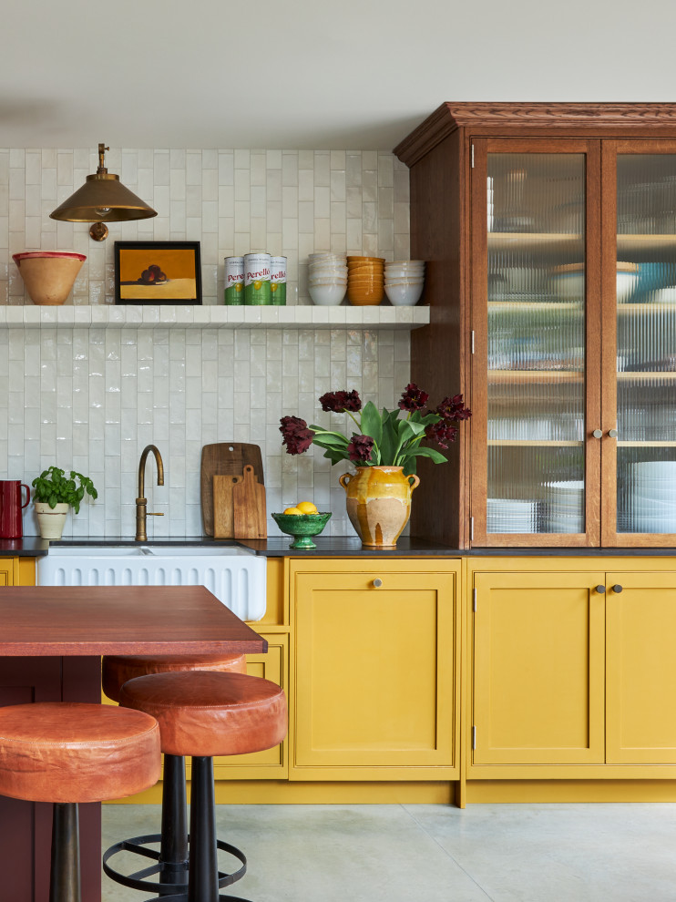 Mid-sized transitional single-wall open concept kitchen photo in London with a farmhouse sink, shaker cabinets, yellow cabinets, solid surface countertops, white backsplash, ceramic backsplash, an island and black countertops