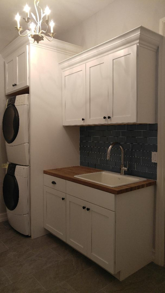 Photo of a mid-sized traditional single-wall dedicated laundry room in Miami with a drop-in sink, shaker cabinets, white cabinets, wood benchtops, grey walls, a stacked washer and dryer, brown benchtop, blue splashback, glass tile splashback, porcelain floors and grey floor.