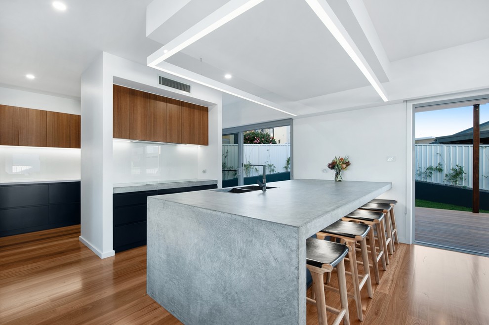 This is an example of a large modern galley kitchen pantry in Newcastle - Maitland with a drop-in sink, flat-panel cabinets, black cabinets, concrete benchtops, white splashback, glass sheet splashback, black appliances, medium hardwood floors, with island, brown floor and grey benchtop.