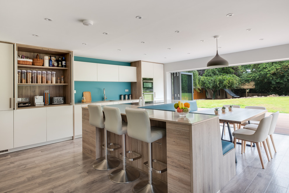 This is an example of a large scandinavian eat-in kitchen in Gloucestershire with flat-panel cabinets, white cabinets, solid surface benchtops, blue splashback, glass sheet splashback, black appliances, porcelain floors, with island, an undermount sink, beige floor and beige benchtop.