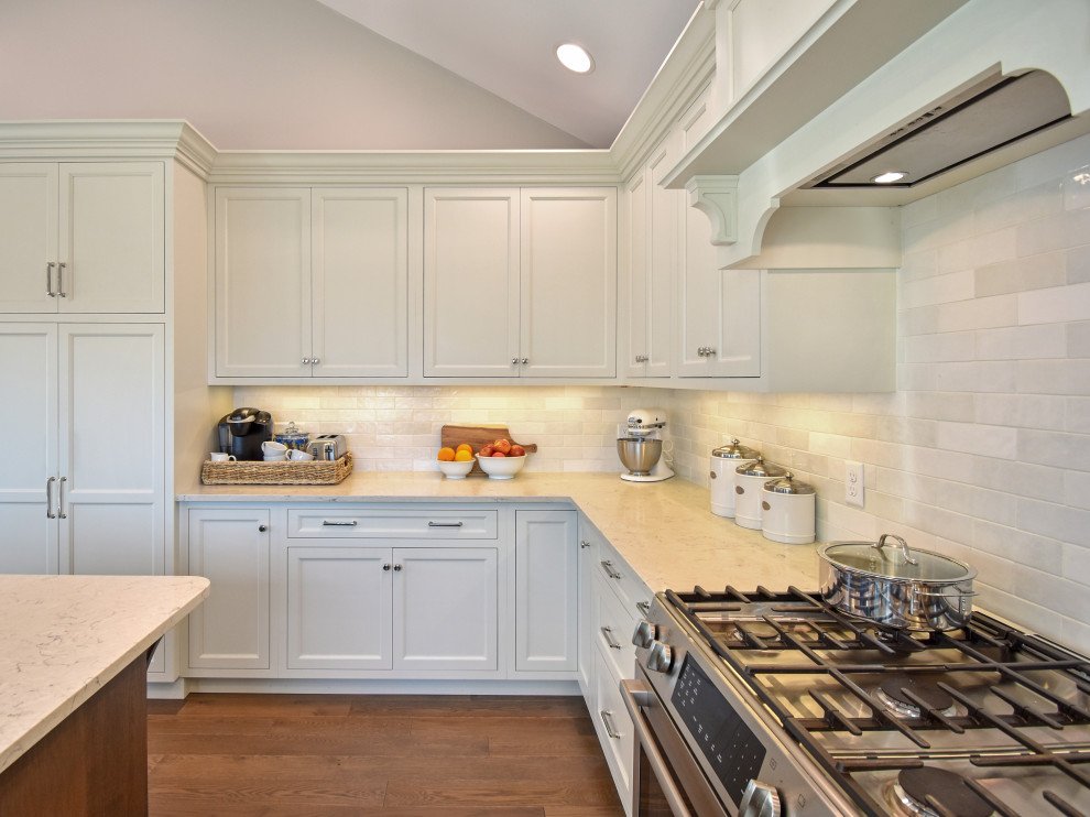 Example of a transitional medium tone wood floor, brown floor and vaulted ceiling eat-in kitchen design with shaker cabinets, white cabinets, quartz countertops, white backsplash, ceramic backsplash, white appliances, an island and white countertops