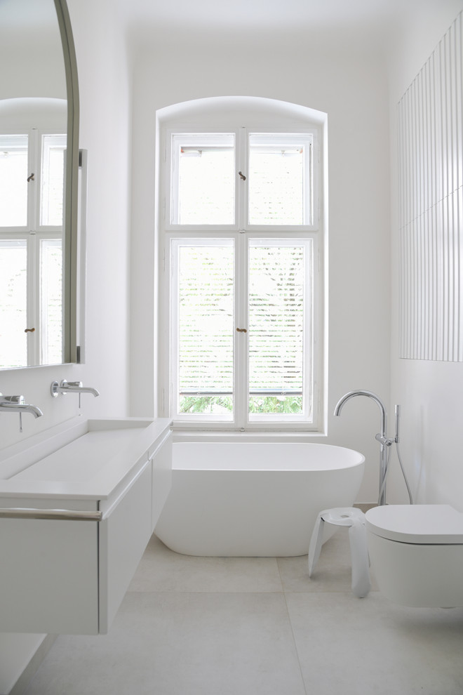 Photo of a mid-sized contemporary master bathroom in Berlin with white cabinets, a freestanding tub, a curbless shower, a two-piece toilet, gray tile, ceramic tile, white walls, a wall-mount sink, solid surface benchtops, a hinged shower door, white benchtops, a double vanity, a floating vanity and flat-panel cabinets.