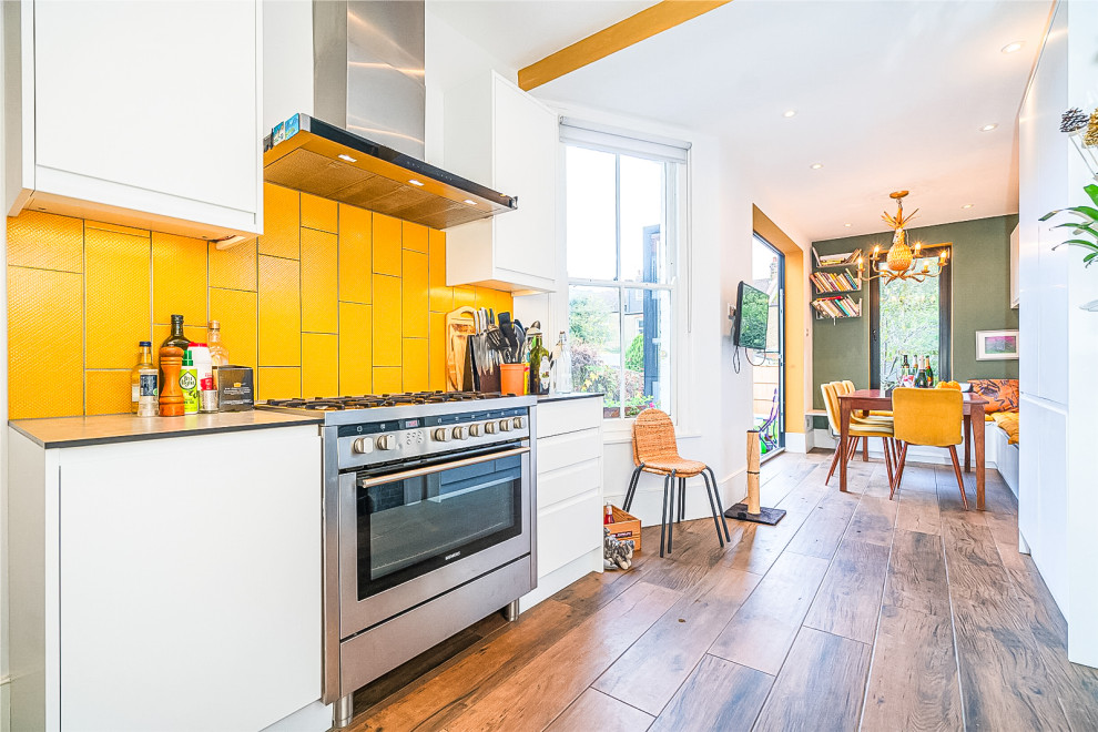 Photo of a small contemporary kitchen/diner in London with flat-panel cabinets, white cabinets, composite countertops, yellow splashback, porcelain splashback, stainless steel appliances, porcelain flooring, no island, brown floors, grey worktops and a chimney breast.