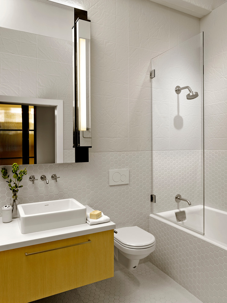 This is an example of a mid-sized industrial bathroom in San Francisco with a vessel sink, flat-panel cabinets, an alcove tub, a shower/bathtub combo, a wall-mount toilet, white tile, white walls and yellow cabinets.