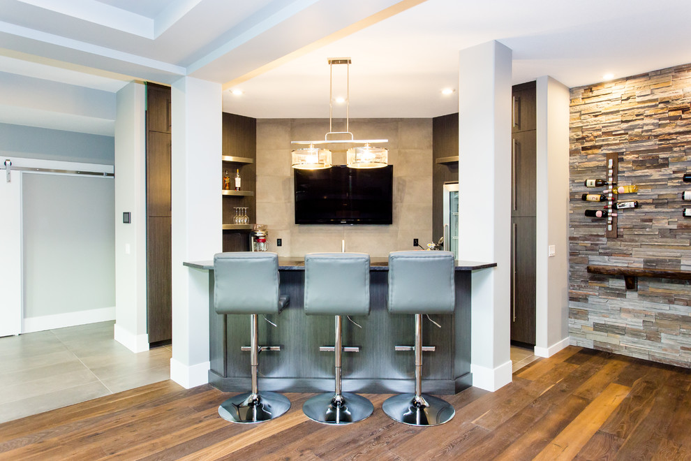 Photo of a large contemporary u-shaped seated home bar in Calgary with an undermount sink, flat-panel cabinets, brown cabinets, granite benchtops, grey splashback, cement tile splashback, vinyl floors and brown floor.