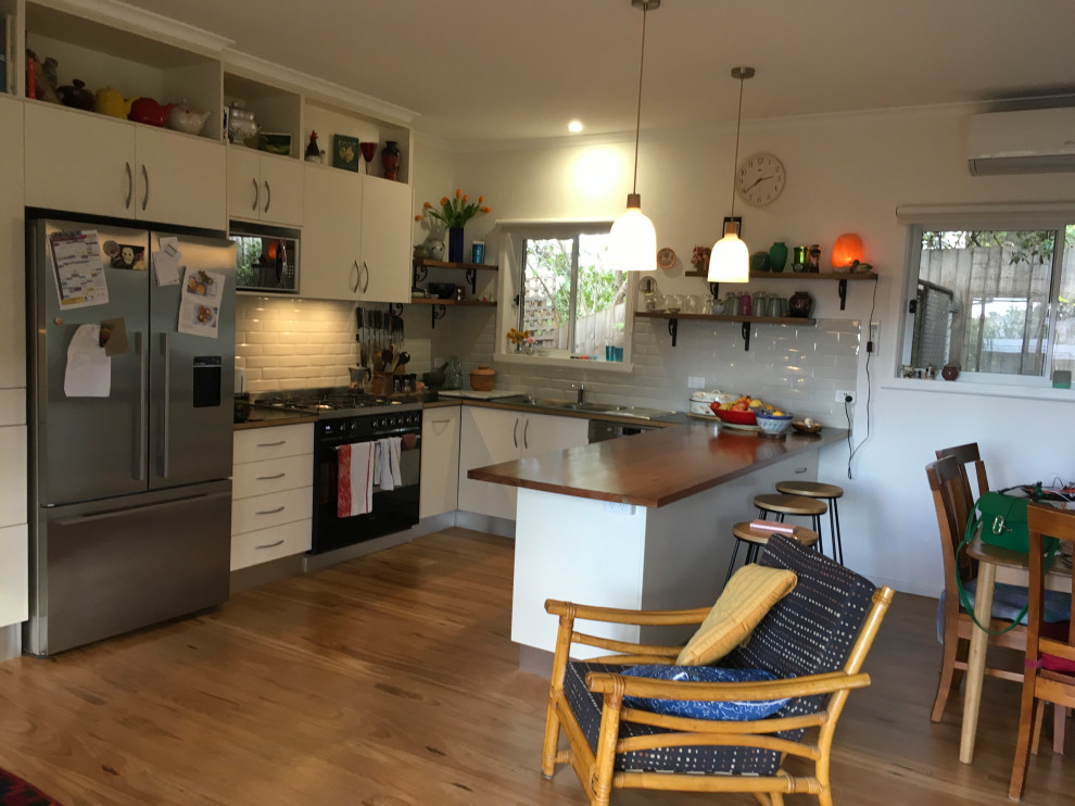 Design ideas for a mid-sized eclectic u-shaped open plan kitchen in Melbourne with a double-bowl sink, flat-panel cabinets, white cabinets, wood benchtops, white splashback, ceramic splashback, stainless steel appliances, medium hardwood floors, a peninsula, brown floor and black benchtop.