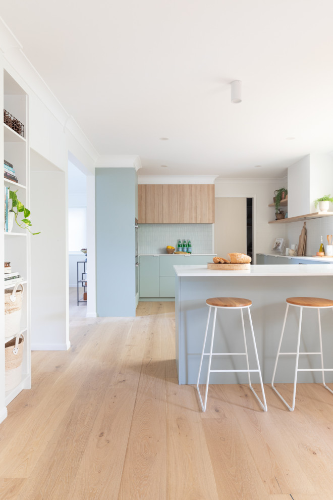 Mid-sized beach style l-shaped kitchen pantry in Brisbane with an undermount sink, flat-panel cabinets, green cabinets, quartz benchtops, blue splashback, mosaic tile splashback, stainless steel appliances, light hardwood floors, a peninsula, beige floor and white benchtop.