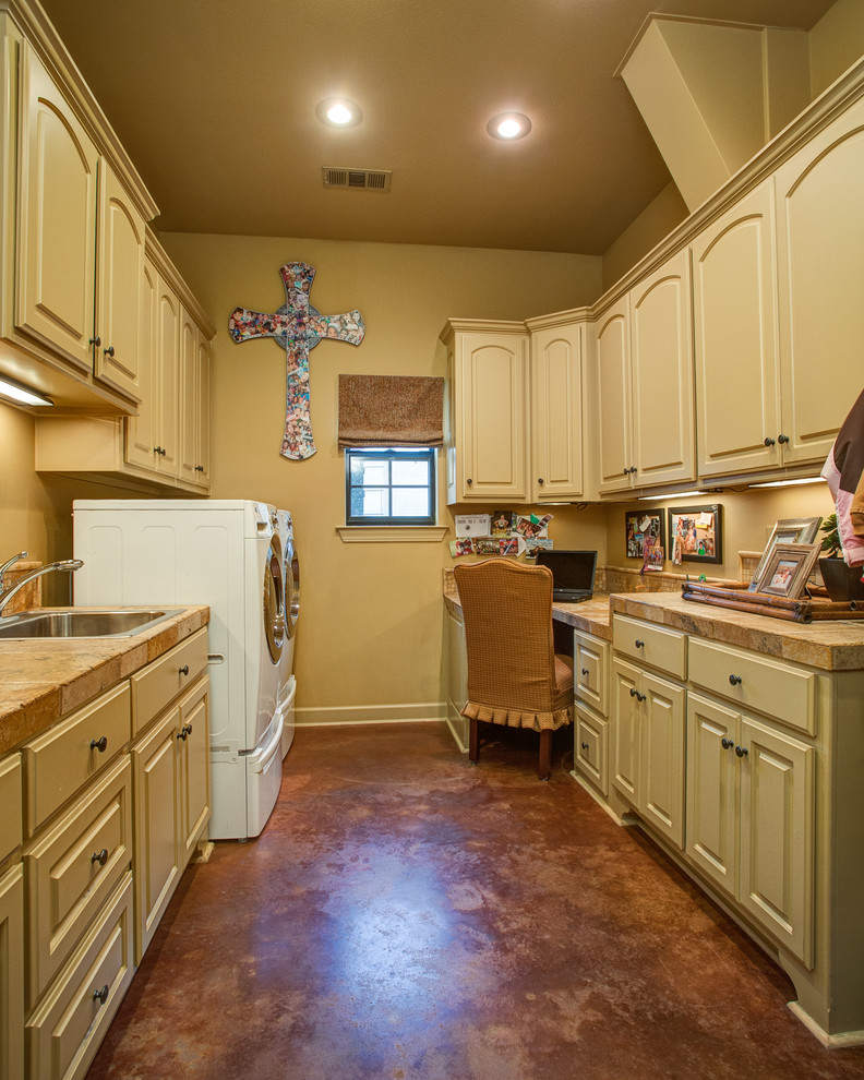 Photo of a large traditional galley utility room in Little Rock with a drop-in sink, raised-panel cabinets, beige cabinets, wood benchtops, beige walls, concrete floors, a side-by-side washer and dryer, brown floor and beige benchtop.