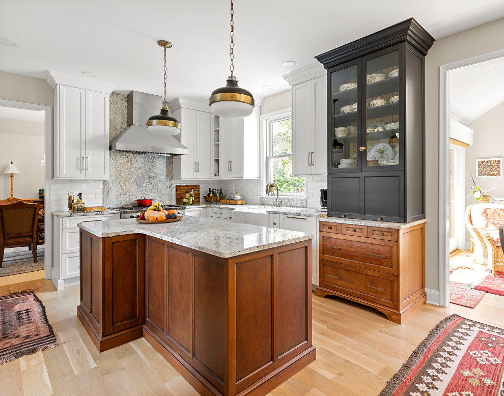 Example of a mid-sized classic l-shaped light wood floor and beige floor enclosed kitchen design in Philadelphia with a farmhouse sink, recessed-panel cabinets, granite countertops, white backsplash, ceramic backsplash, stainless steel appliances, an island and multicolored countertops