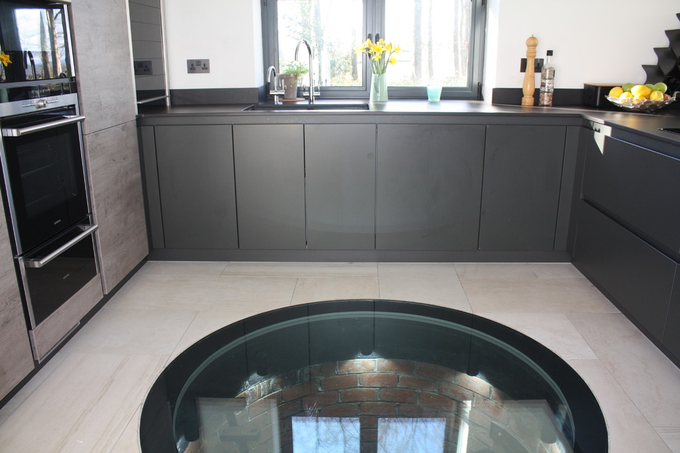Photo of a mid-sized modern u-shaped open plan kitchen in Other with a peninsula, an undermount sink, flat-panel cabinets, grey cabinets, solid surface benchtops, grey splashback, black appliances, ceramic floors, engineered quartz splashback, grey floor, grey benchtop and coffered.