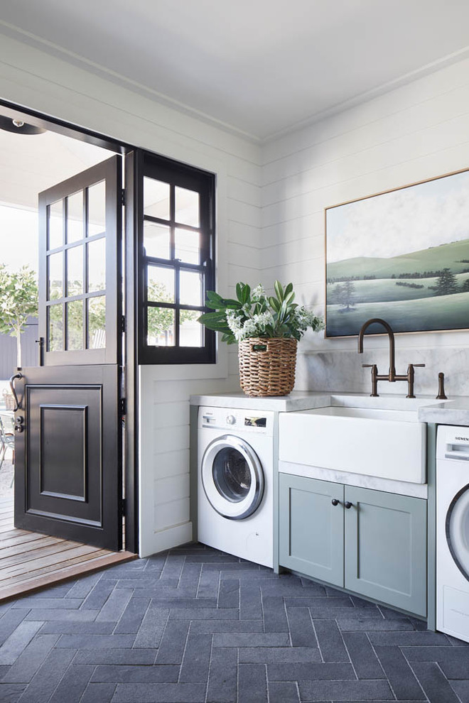 Photo of a transitional single-wall laundry room in Sydney with a farmhouse sink, shaker cabinets, grey cabinets, white walls, a side-by-side washer and dryer, black floor and white benchtop.