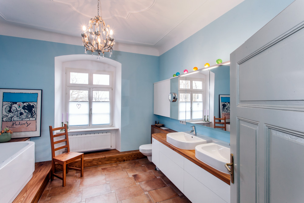 Inspiration for a mid-sized country bathroom in Stuttgart with white cabinets, wood benchtops, brown benchtops, a wall-mount toilet, brown tile, blue walls, terra-cotta floors, brown floor, flat-panel cabinets, an alcove tub, a corner shower, a drop-in sink and a hinged shower door.