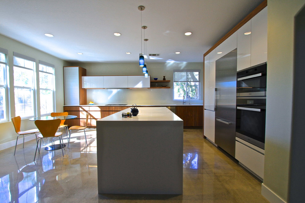 Mid-sized contemporary l-shaped eat-in kitchen in Orange County with an undermount sink, flat-panel cabinets, white cabinets, solid surface benchtops, white splashback, glass sheet splashback, stainless steel appliances, concrete floors and with island.