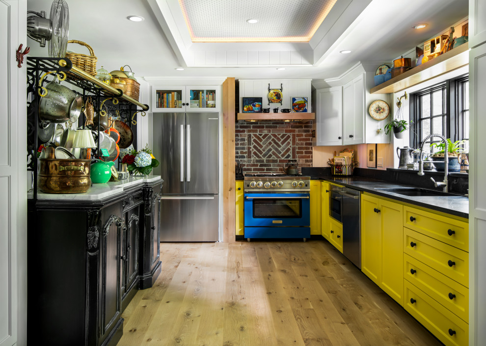 Foto på ett mellanstort vintage svart kök, med en undermonterad diskho, skåp i shakerstil, gula skåp, bänkskiva i täljsten, svart stänkskydd, stänkskydd i trä, färgglada vitvaror, ljust trägolv och brunt golv