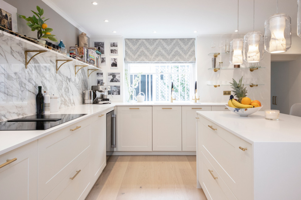 This is an example of a medium sized traditional u-shaped kitchen/diner in Oxfordshire with a double-bowl sink, shaker cabinets, white cabinets, composite countertops, white splashback, marble splashback, black appliances, light hardwood flooring, an island, brown floors, white worktops and feature lighting.